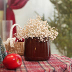 A glossy dark brown ceramic vase filled with dried baby's breath flowers, set on a red plaid tablecloth with a wicker basket, a red apple, and a blurred festive background.