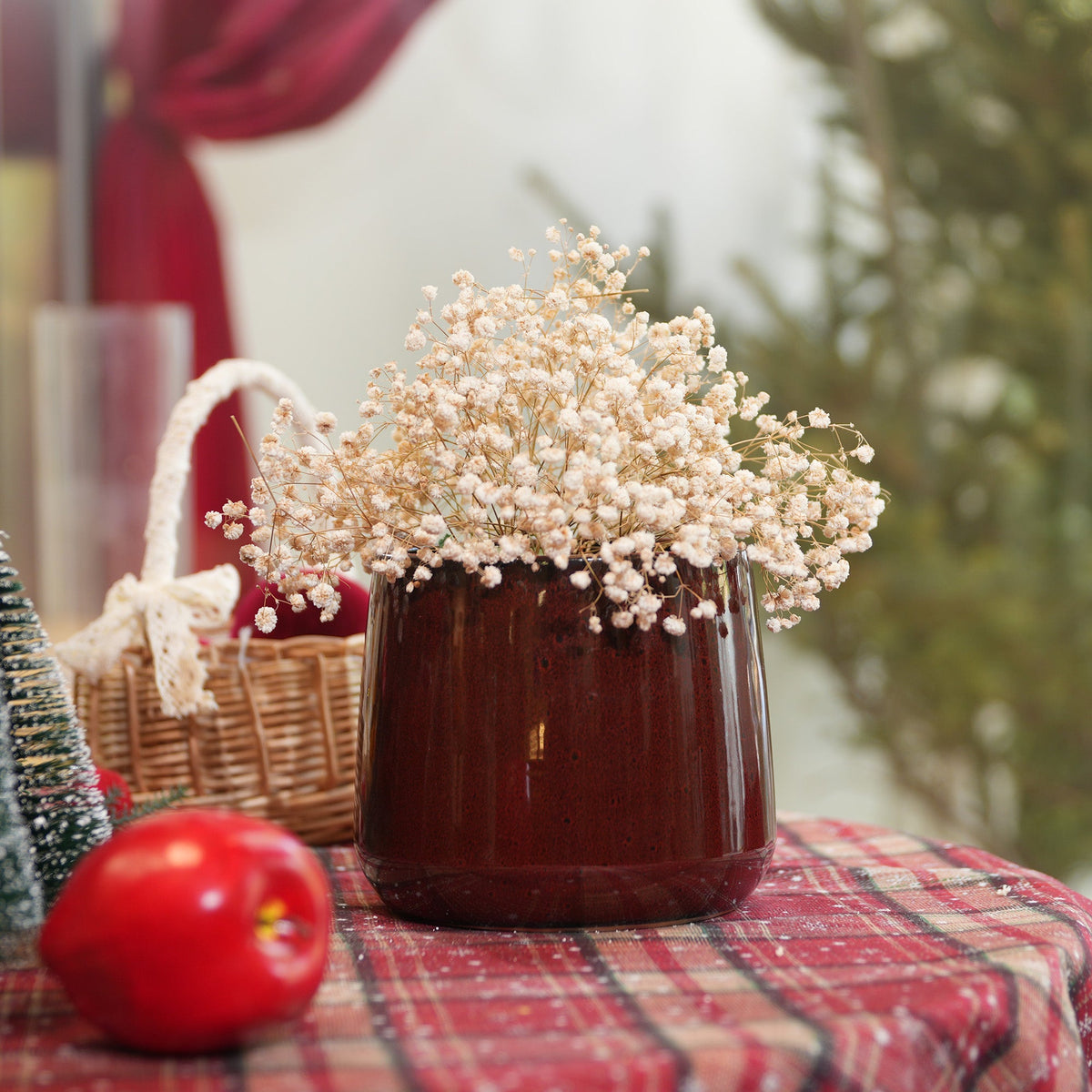 A glossy dark brown ceramic vase filled with dried baby's breath flowers, set on a red plaid tablecloth with a wicker basket, a red apple, and a blurred festive background.