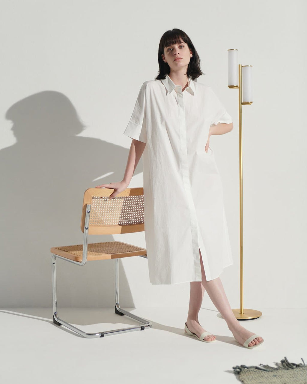 Woman wearing a white dress standing in a minimalistic room with a chair and floor lamp.