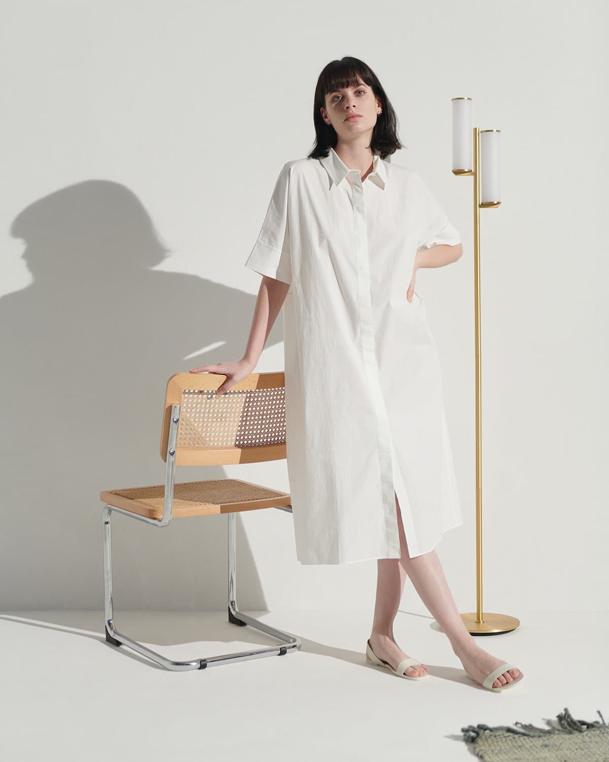 Woman wearing a white dress standing in a minimalistic room with a chair and floor lamp.