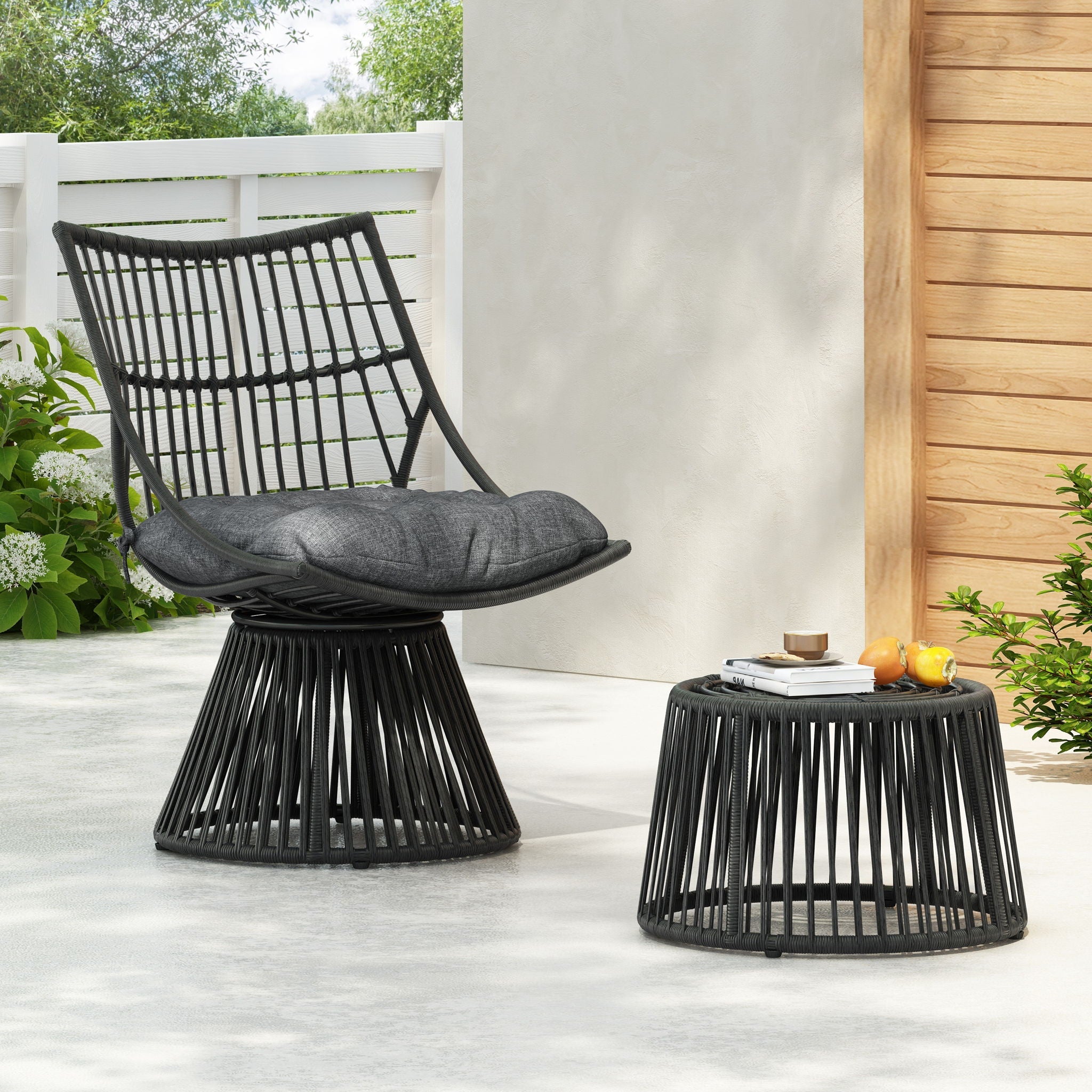 Black wicker chair and side table on a patio with a wooden wall and plants in the background.