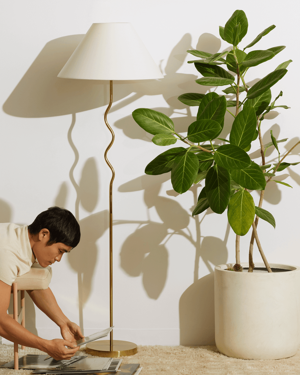 Person sitting on a stool reading a book next to a floor lamp and potted plant.