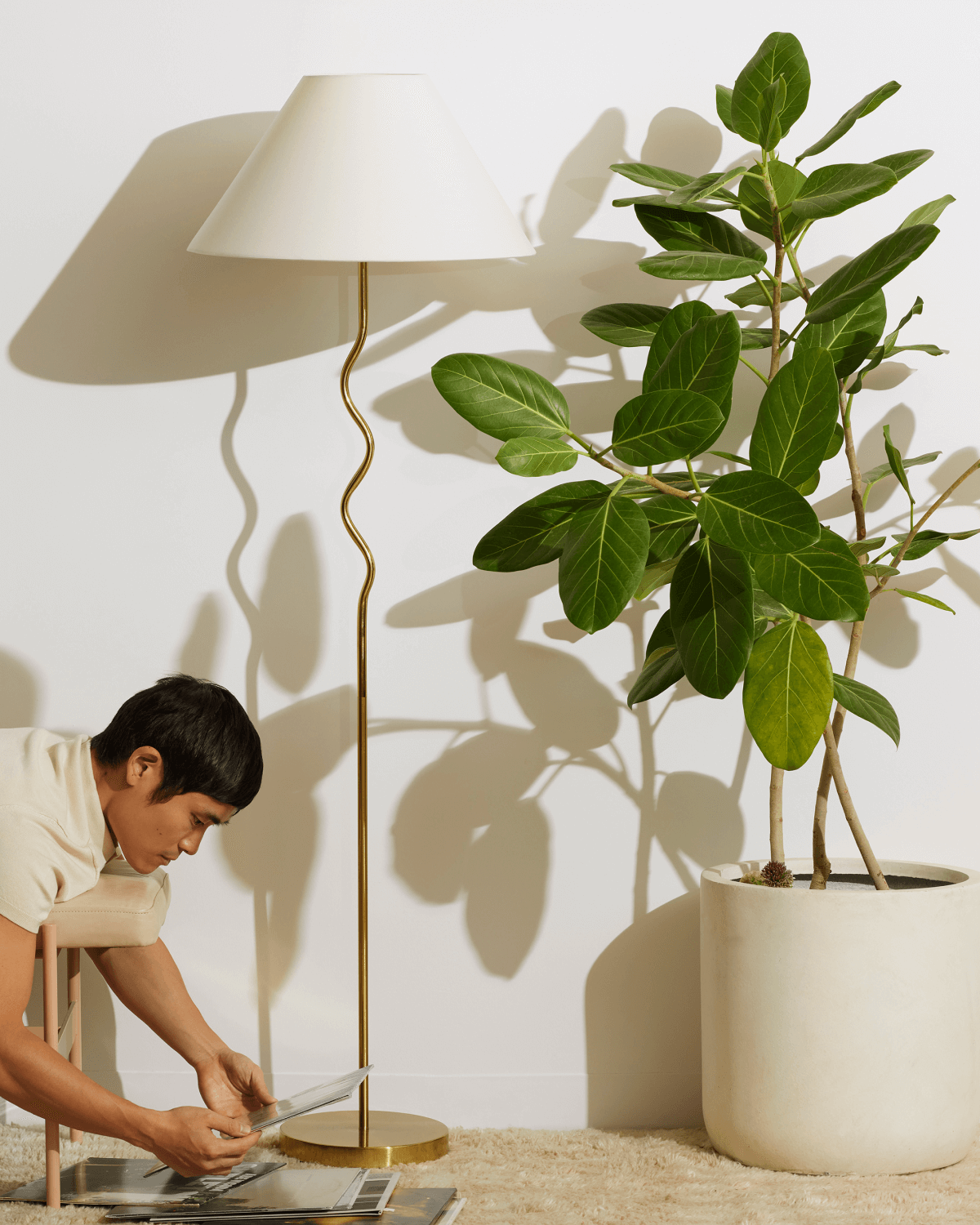 Person sitting on a stool reading a book next to a floor lamp and potted plant.