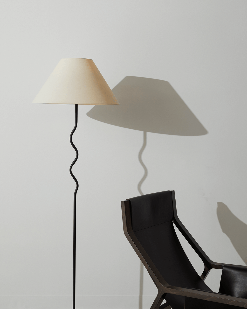 Floor lamp with a beige shade casting a shadow on a white wall next to a black chair.
