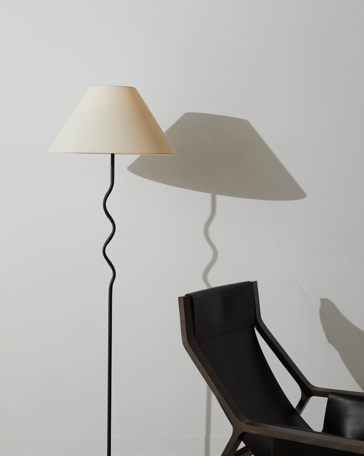 Floor lamp with a beige shade casting a shadow on a white wall next to a black chair.
