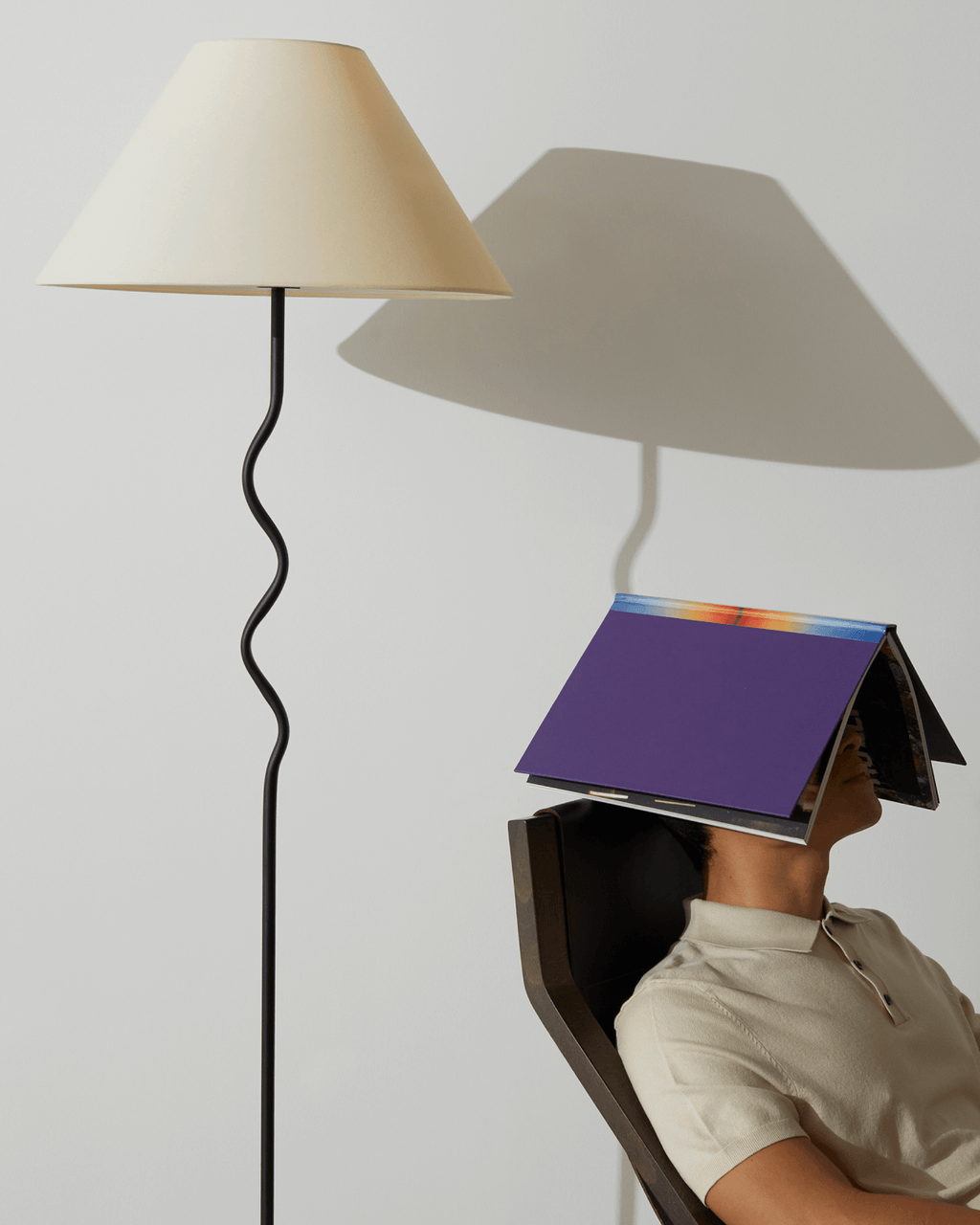 Floor lamp with a beige shade casting a shadow on a white wall next to a man napping on a black chair with a book on his head.