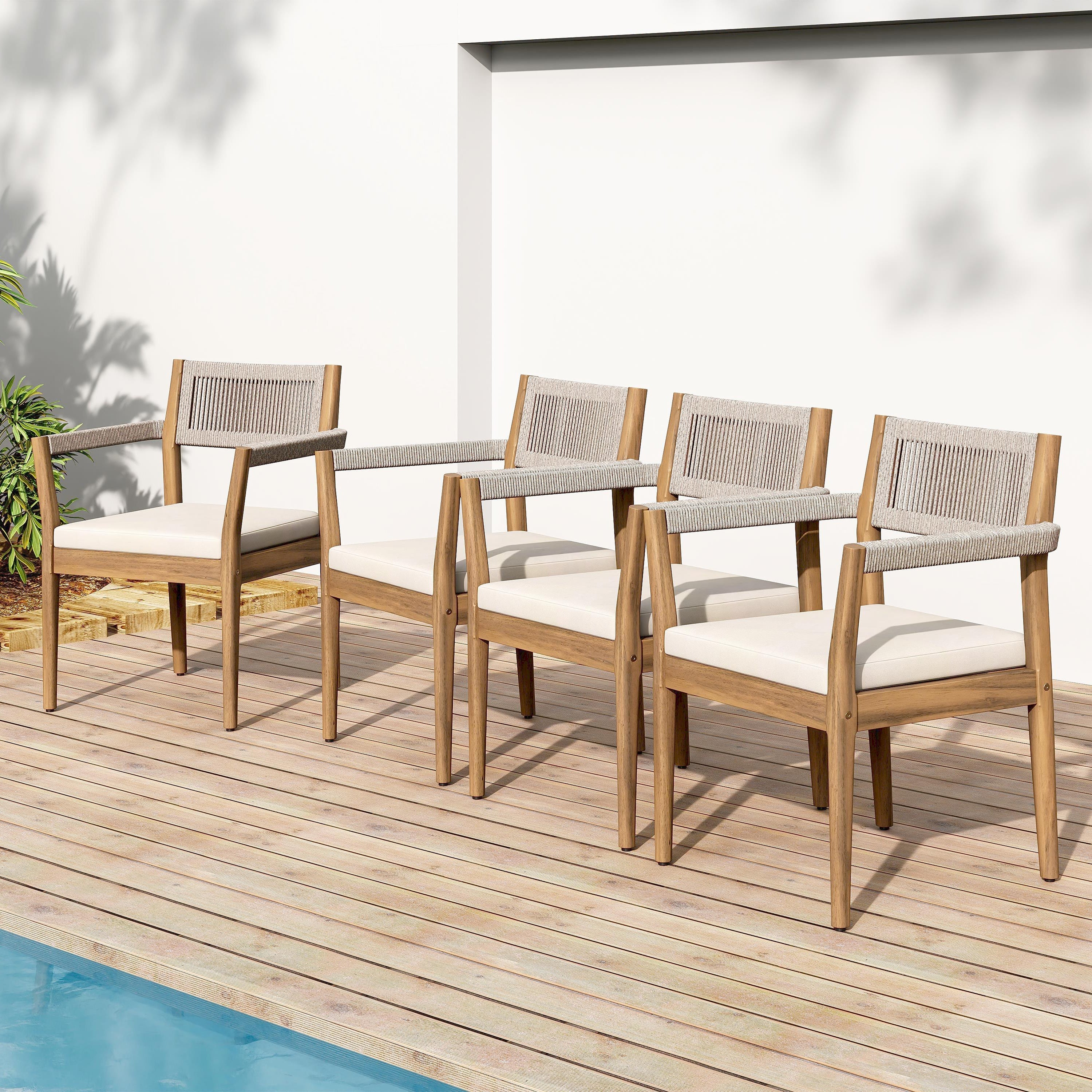 Set of four wooden chairs with white cushions on a wooden deck by a pool.