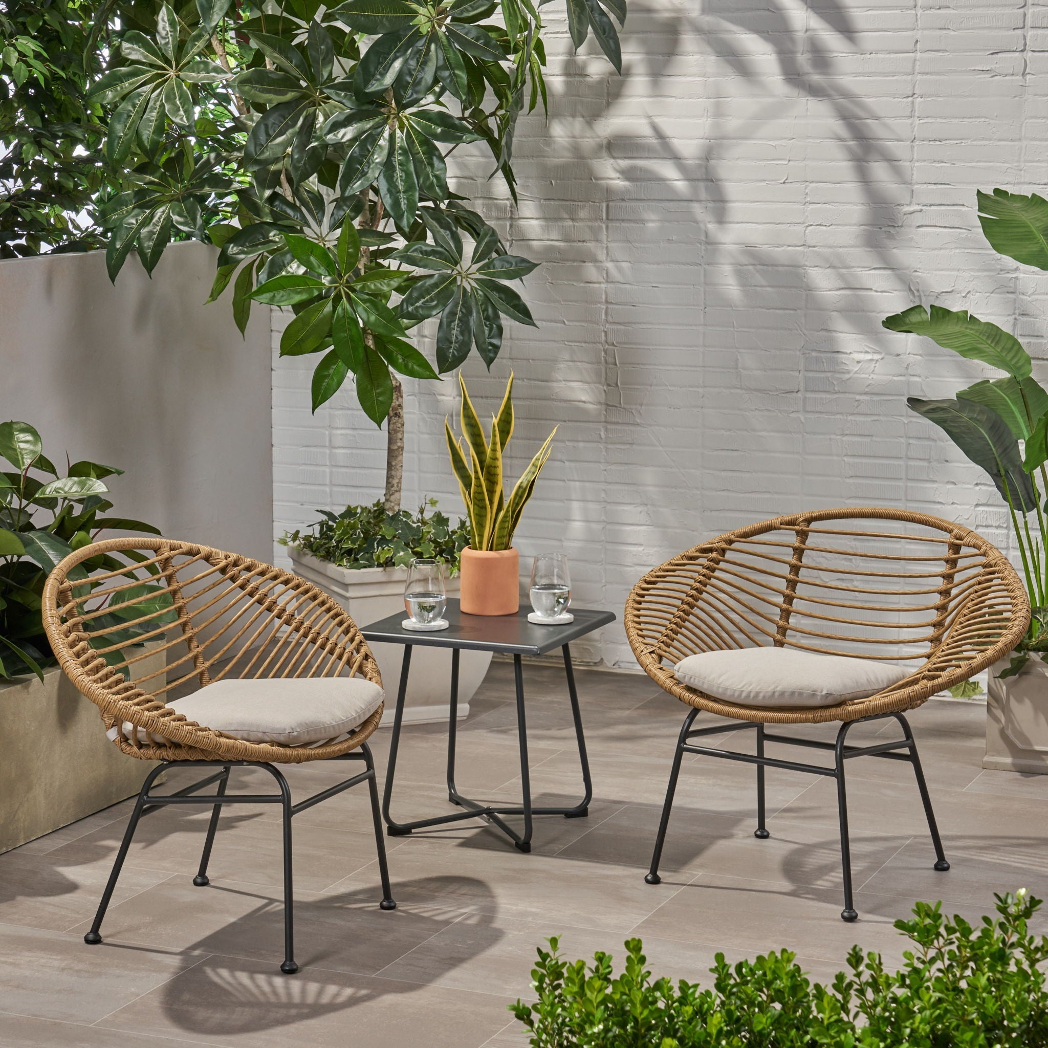 Two wicker chairs with cushions and a small table on a patio with plants and a white wall.