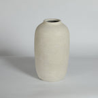 A minimalist ceramic vase with a rounded, bulbous shape and a short, slightly flared neck, featuring a textured matte finish in a warm off-white or cream color. It is photographed against a neutral light gray studio background.