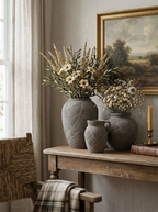 Three textured gray ceramic vases of different sizes arranged on a rustic wooden console table, filled with dried grasses, white daisies, and baby's breath flowers. The setting is a cozy room with a beige wall, a framed landscape painting, a window with sheer curtains, and a woven chair.