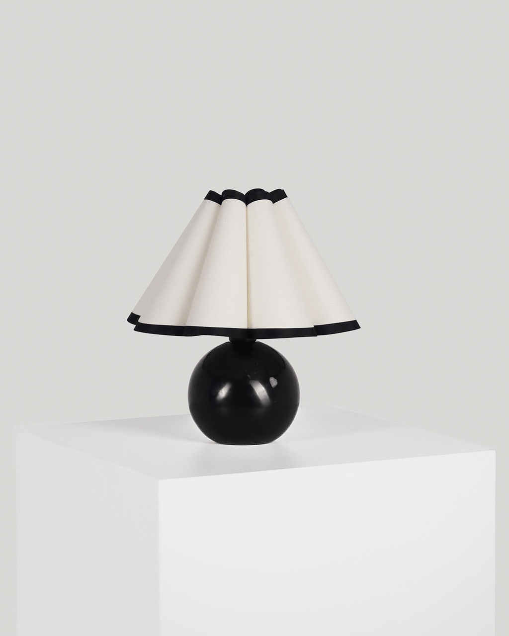 A modern table lamp with a glossy black spherical base and a pleated cream-colored shade trimmed with black, displayed on a white pedestal against a light gray background.