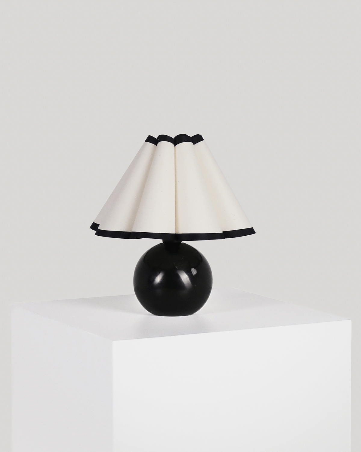 A modern table lamp with a glossy black spherical base and a pleated cream-colored shade trimmed with black, displayed on a white pedestal against a light gray background.
