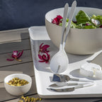 White salad server set on a wooden table with a bowl of salad and flowers.