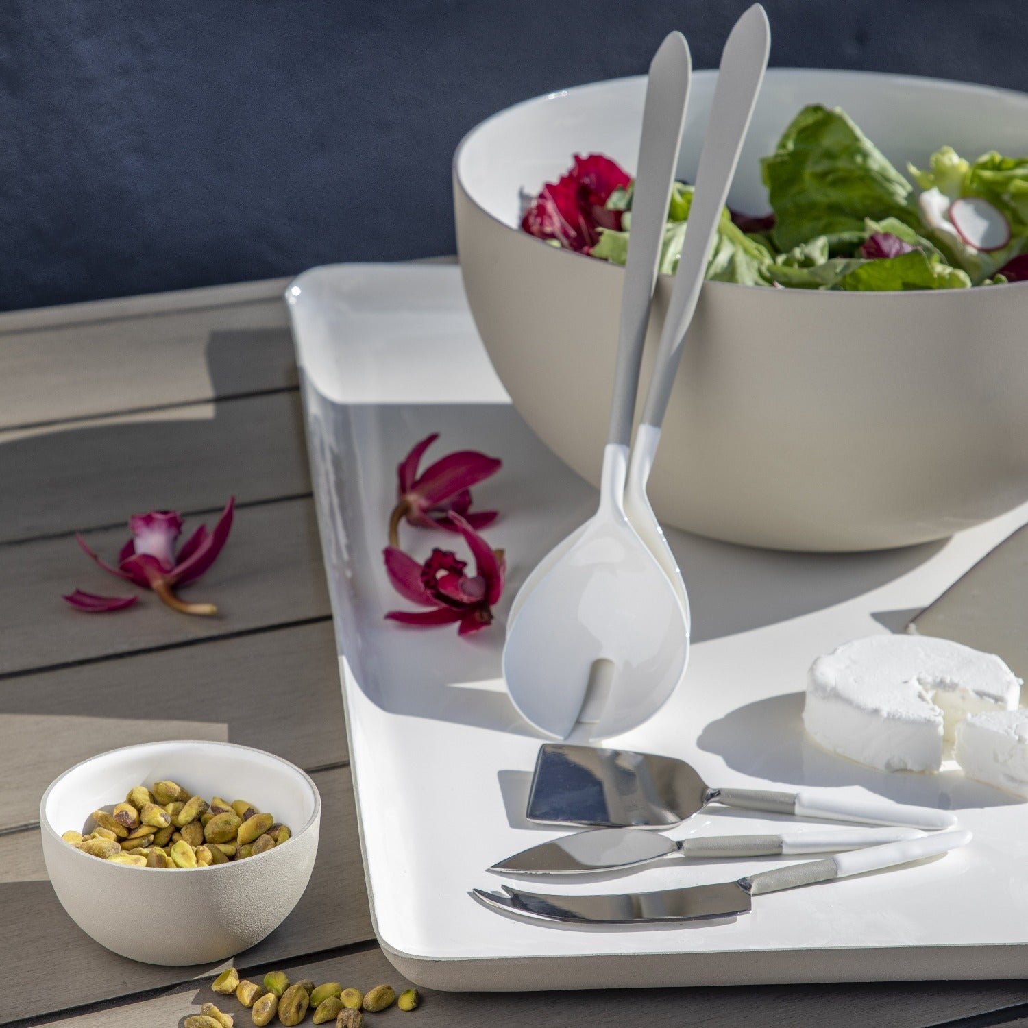White salad server set on a wooden table with a bowl of salad and flowers.