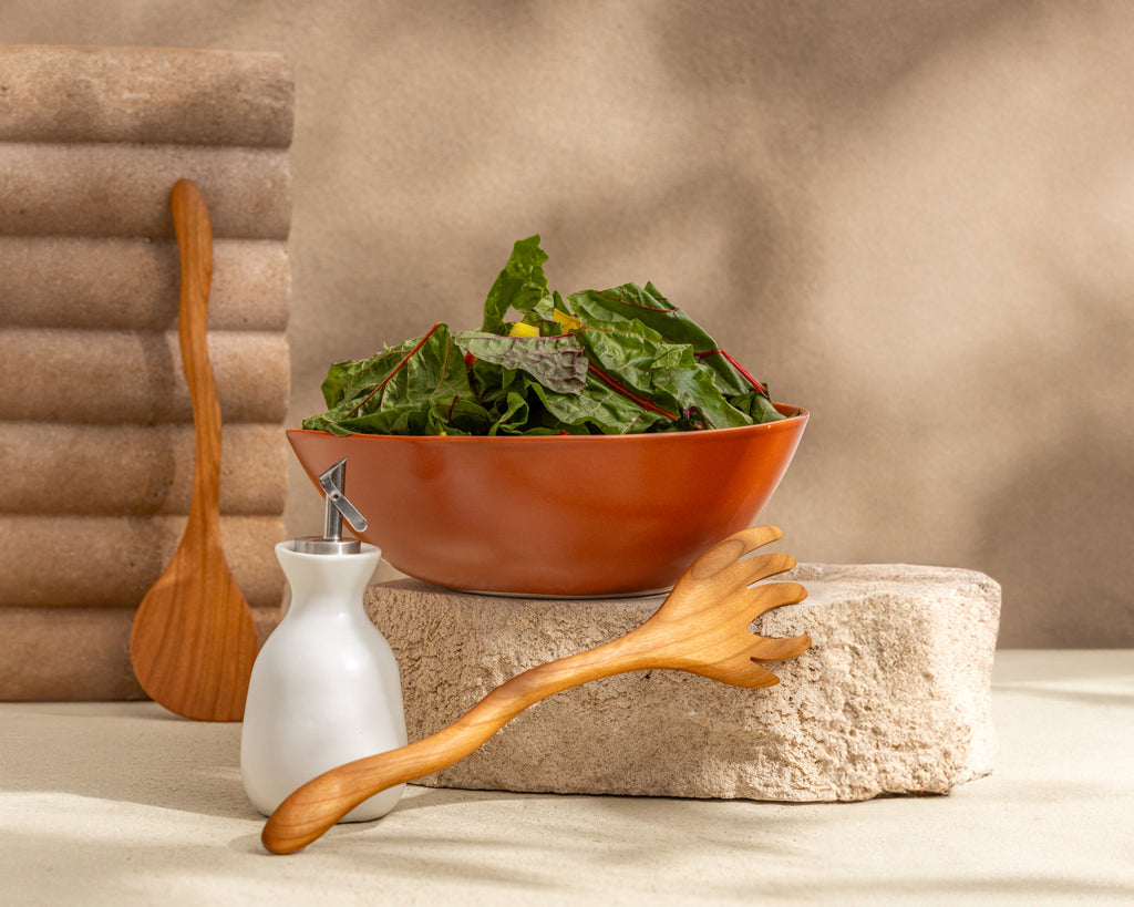 Bowl of greens with a wooden spoon, salt shaker, and wooden spatula on a stone surface.