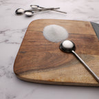 Wooden cutting board with a spoonful of salt and measuring spoons on a marble surface