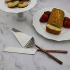 Two cake slices with strawberries on a plate, a stand with cookies, and two cake knives on a marble surface.