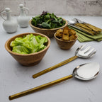 Wooden bowls with salad and croutons, silver and gold utensils on a light surface.