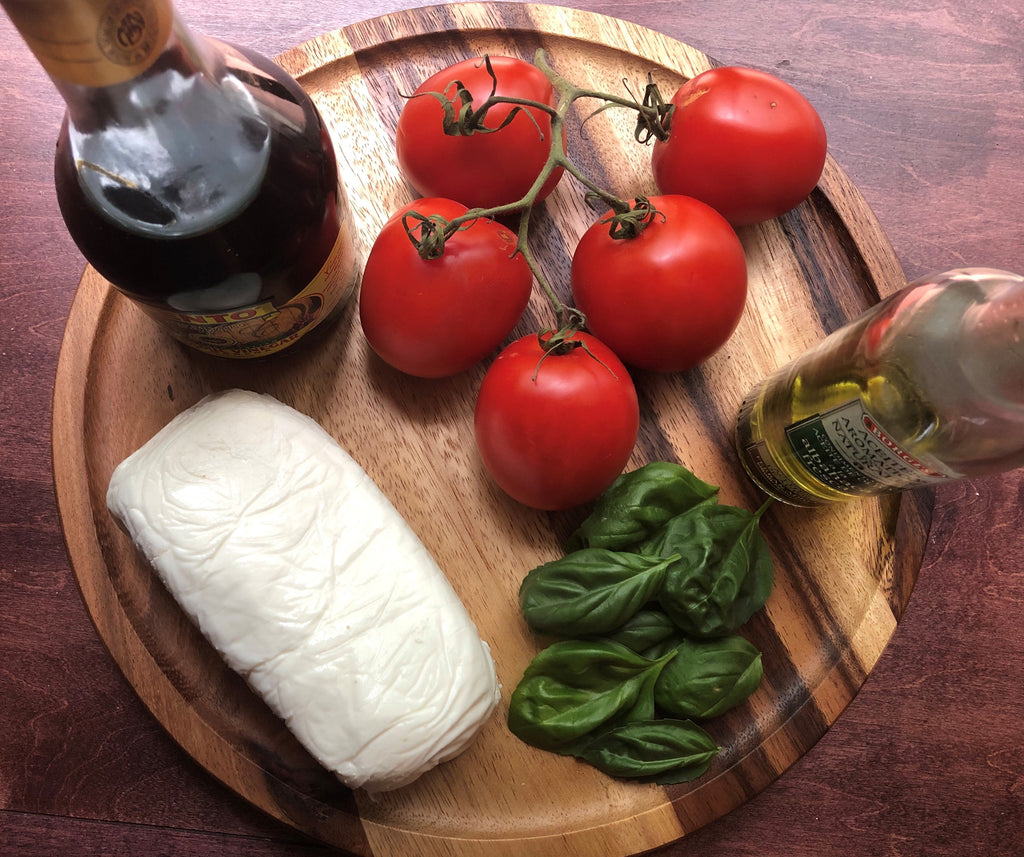 Wooden cutting board with tomatoes, basil, and a block of cheese on a wooden surface.