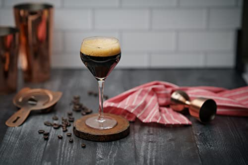 Glass of dark liquid with foam on a wooden coaster, surrounded by coffee beans and a red cloth.