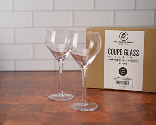 Two coupe glasses on a wooden surface with a branded box in the background.