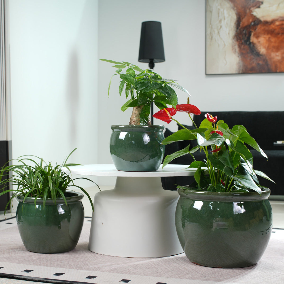 Three glossy dark green ceramic planters arranged on a white pedestal table in a modern living room setting, containing green and red tropical plants.