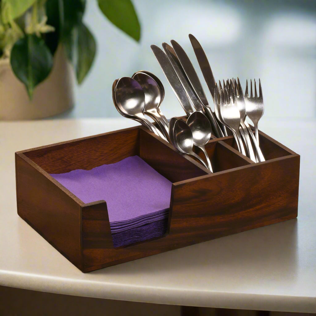 Wooden cutlery holder with silverware and purple napkins on a white surface.