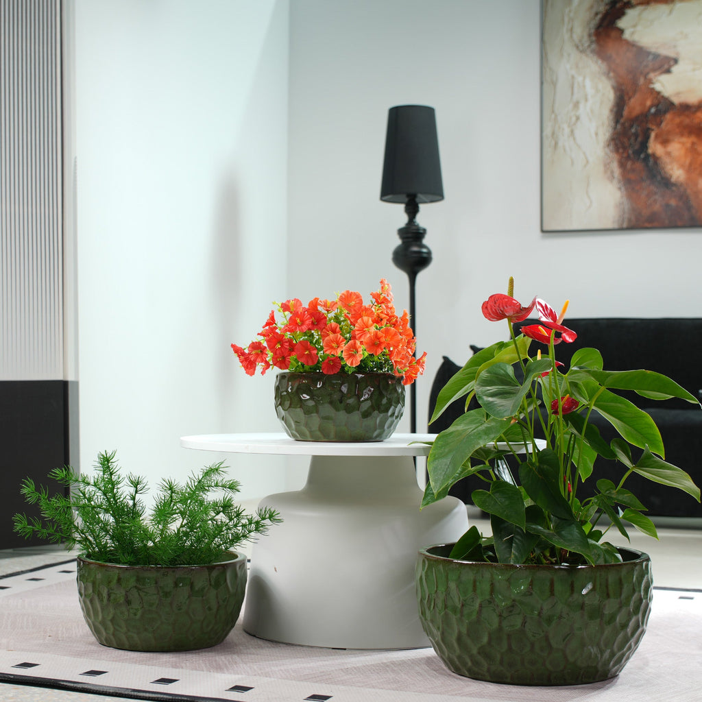 Three textured dark green ceramic planters containing green, orange, and red houseplants, styled on a white pedestal table in a modern living room setting.