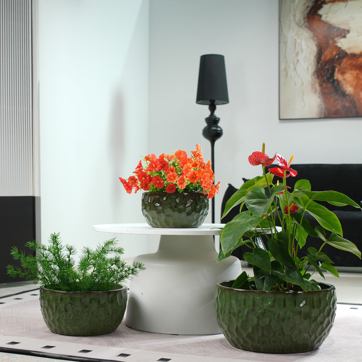 Three textured dark green ceramic planters containing green, orange, and red houseplants, styled on a white pedestal table in a modern living room setting.
