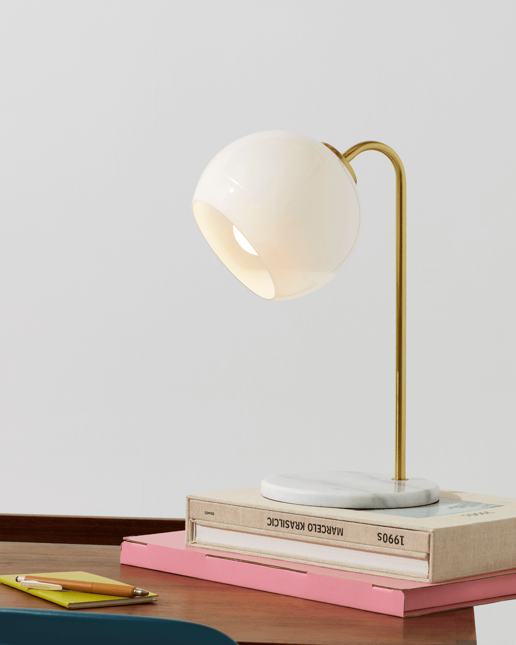 Table lamp with a white shade and gold arm on top of books on a wooden surface.