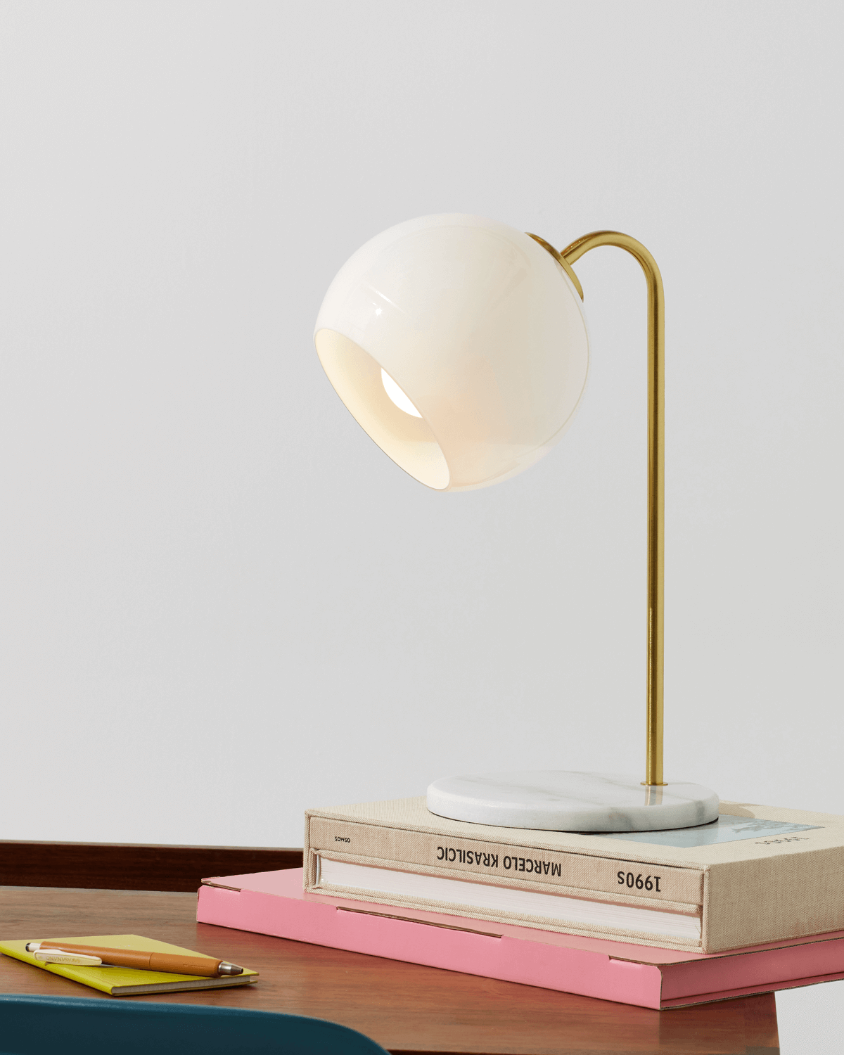 Table lamp with a white shade and gold arm on top of books on a wooden surface.