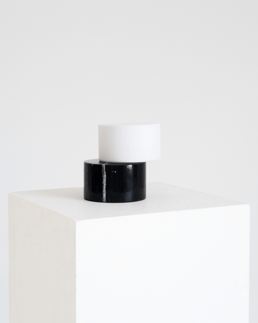 A minimalist two-tier table lamp with a glossy black cylindrical base and a matte white cylindrical shade, displayed on a white cube pedestal against a white background.