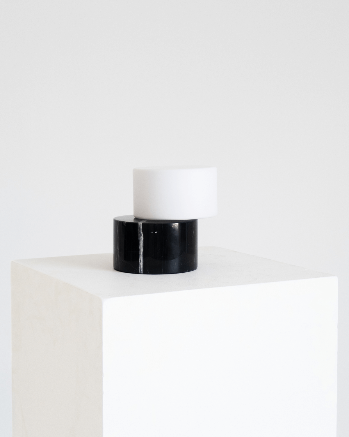 A minimalist two-tier table lamp with a glossy black cylindrical base and a matte white cylindrical shade, displayed on a white cube pedestal against a white background.