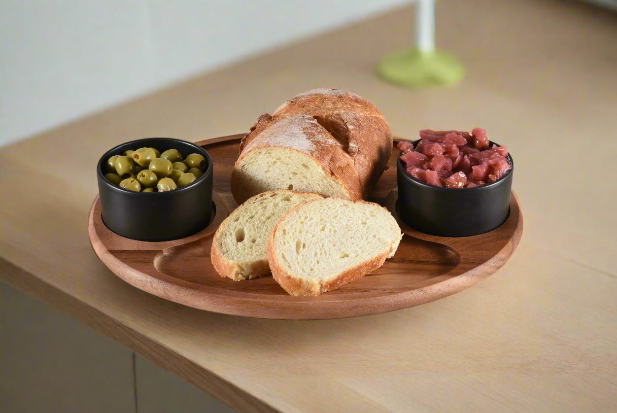 Wooden tray with bread slices, olives, and dried fruits on a light wooden surface.