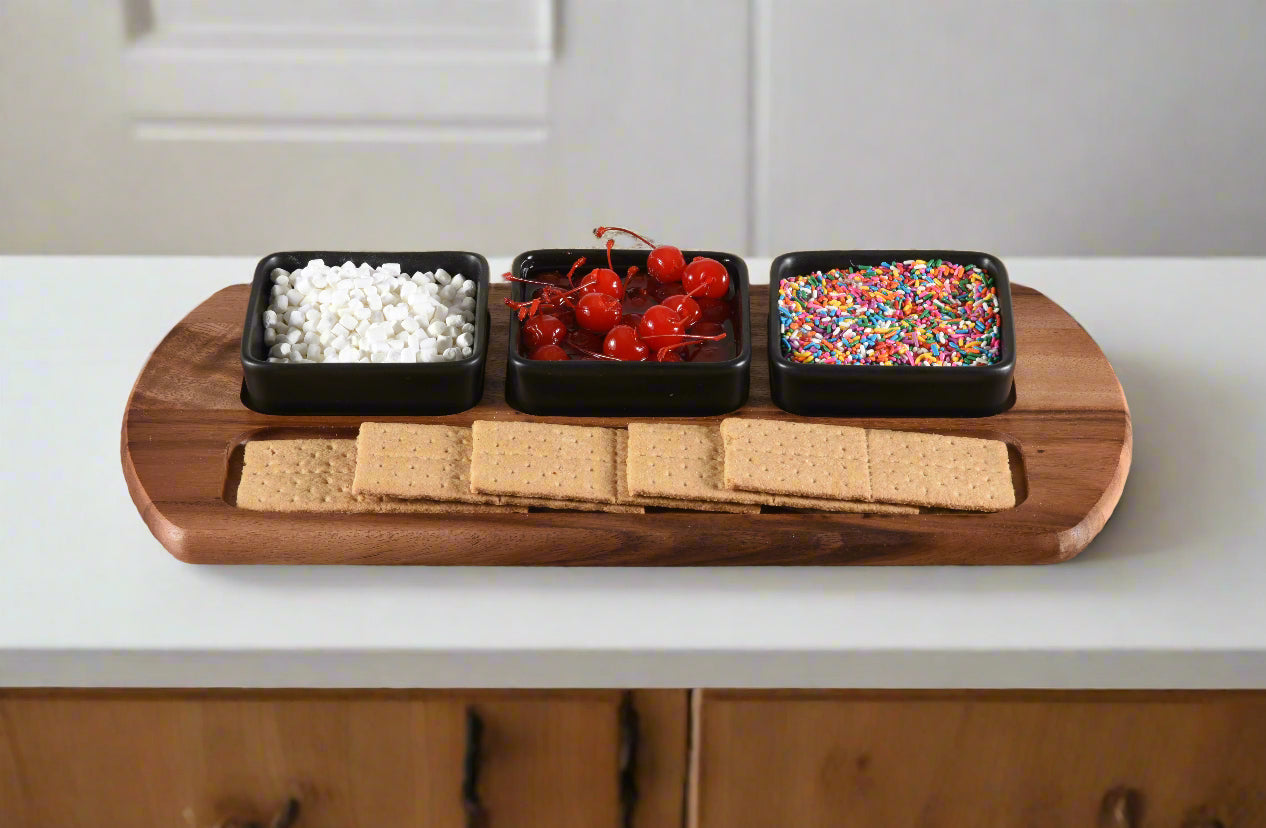 Wooden board with three black containers holding white, red, and colorful sprinkles, along with graham crackers on a kitchen counter.