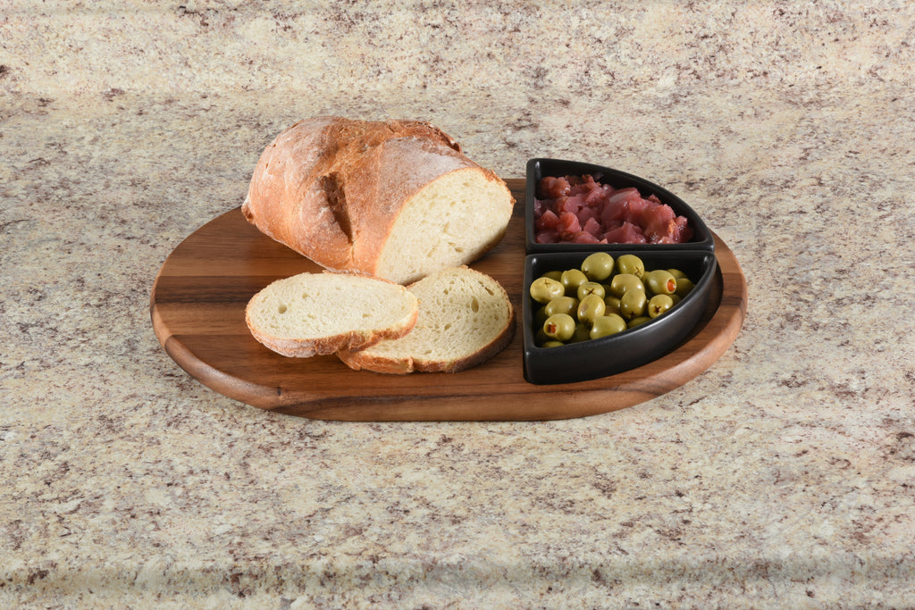 Wooden cutting board with bread slices and a black container of olives and cured meats on a granite countertop.