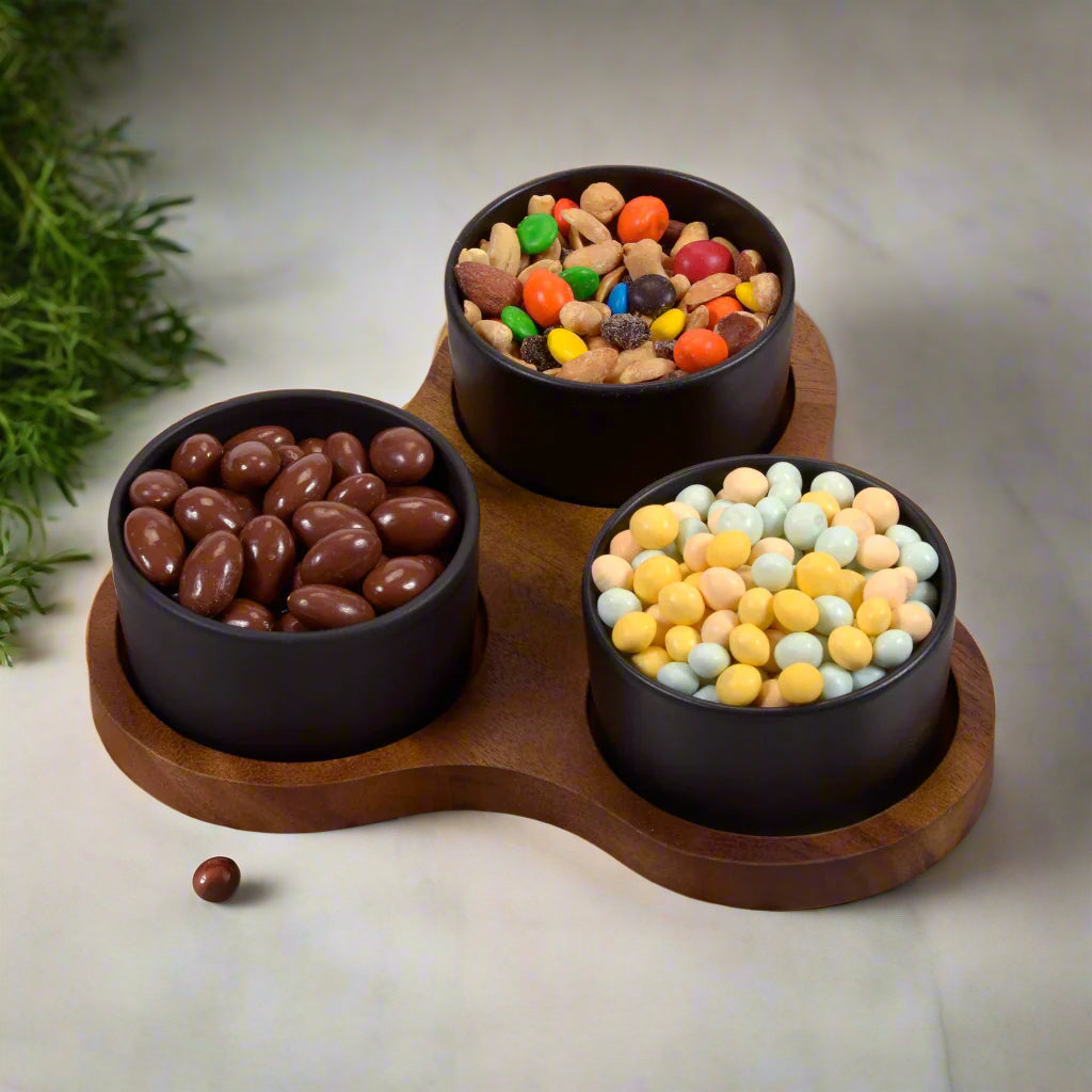 Three black bowls filled with various candies on a wooden tray with a light background