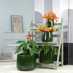 Three glossy dark green ceramic planters arranged on and beside a light gray wooden ladder stand, with orange and yellow flowers placed in the pots, set in a bright indoor room.