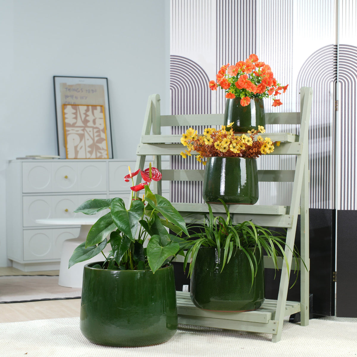 Three glossy dark green ceramic planters arranged on and beside a light gray wooden ladder stand, with orange and yellow flowers placed in the pots, set in a bright indoor room.