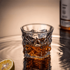Glass of whiskey with a lemon wedge on a reflective surface, with a bottle in the background.