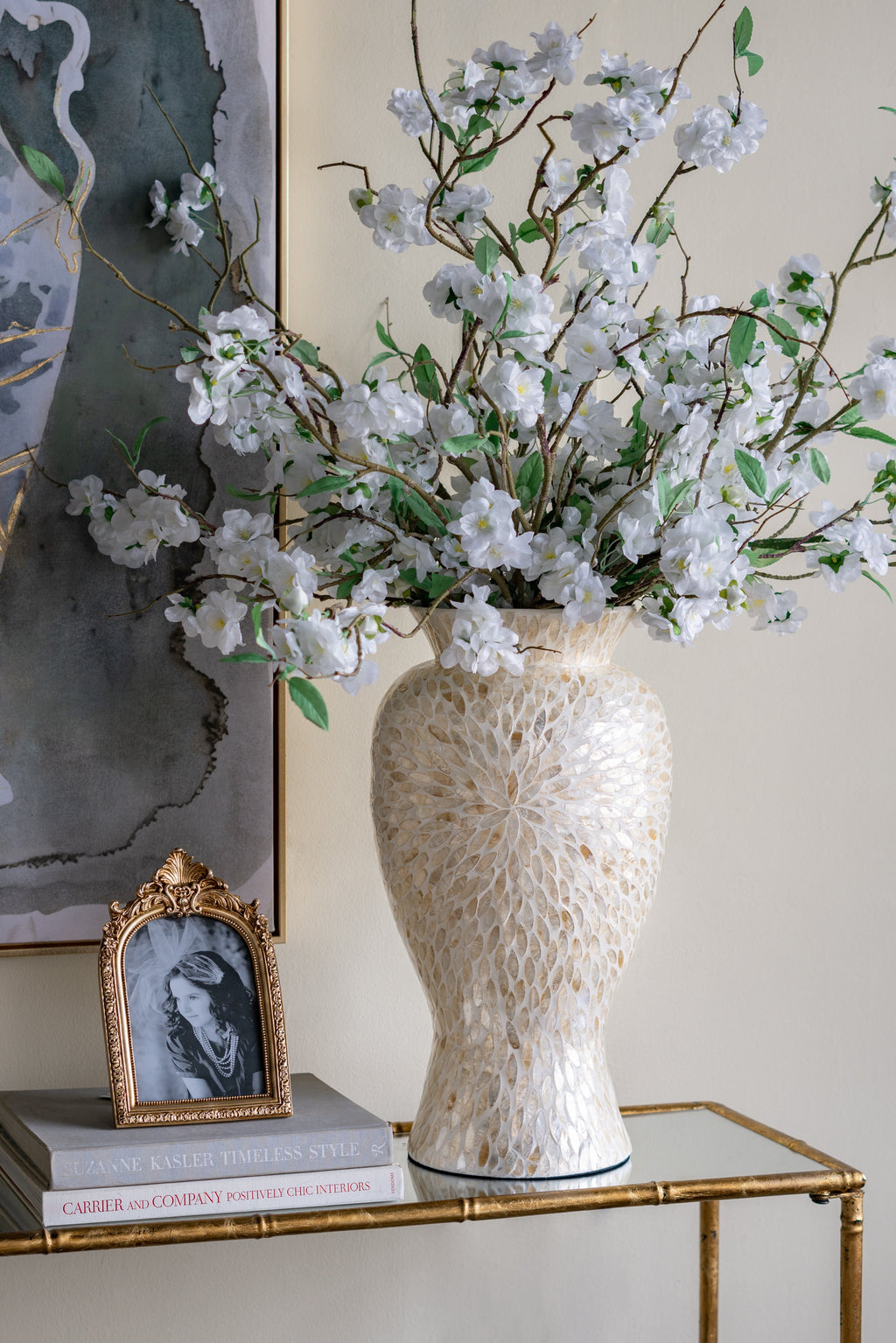 A tall textured cream ceramic vase filled with white cherry blossom branches and green leaves, displayed on a gold-framed console table next to a small framed portrait.