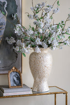 A tall textured cream ceramic vase filled with white cherry blossom branches and green leaves, displayed on a gold-framed console table next to a small framed portrait.