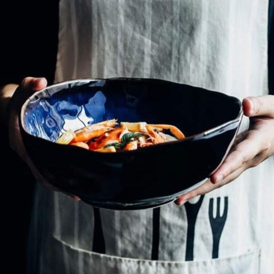 Hands holding large Amelia porcelain soup bowl with pasta — deep cobalt blue glaze, organic irregular rim, bronze edge