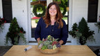 Woman standing behind a table with a decorative plant arrangement in front of a house.