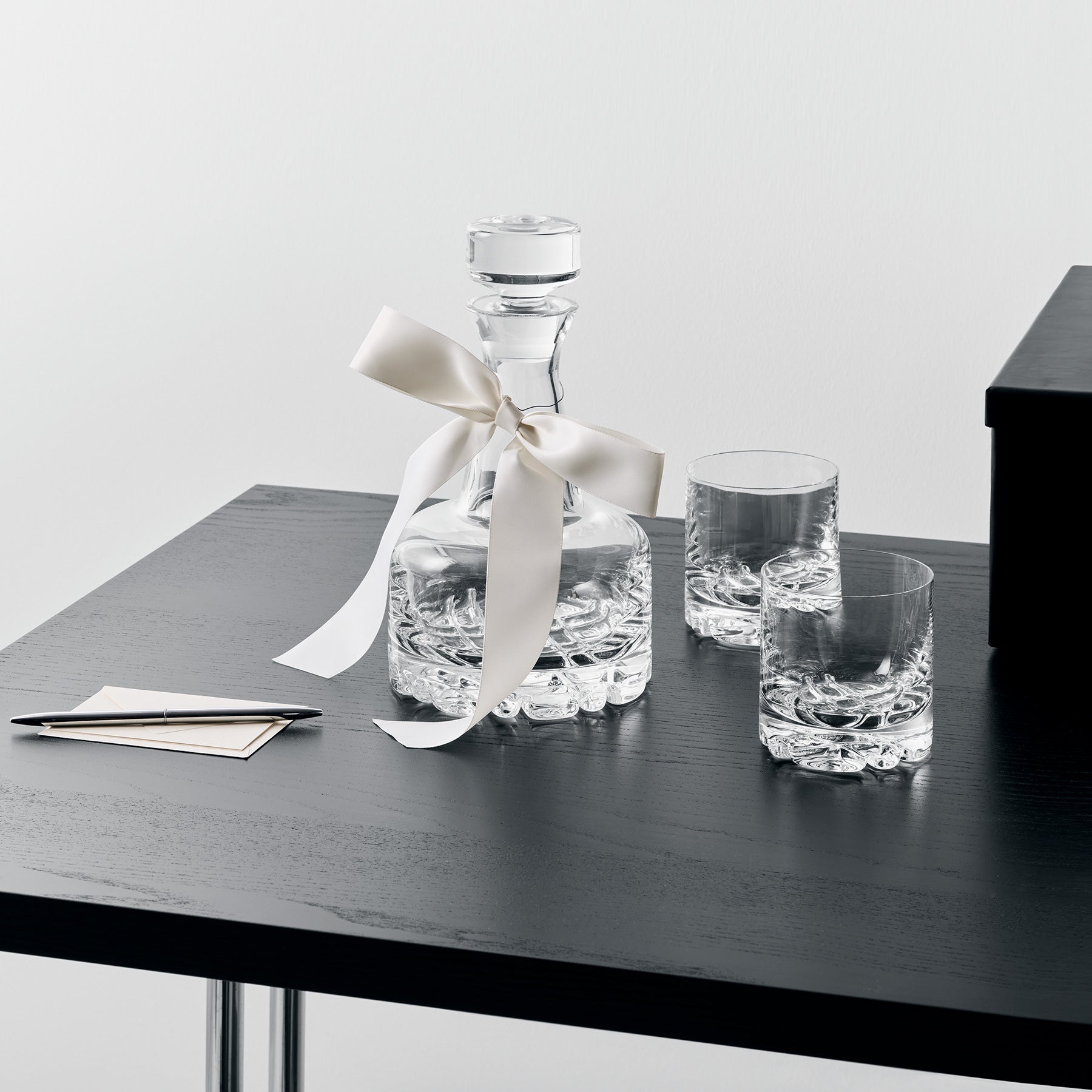Clear glass decanter with a white ribbon and two glasses on a black table.
