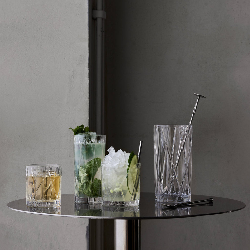 Four cocktails in clear glasses with ice and garnishes on a dark surface against a gray wall.