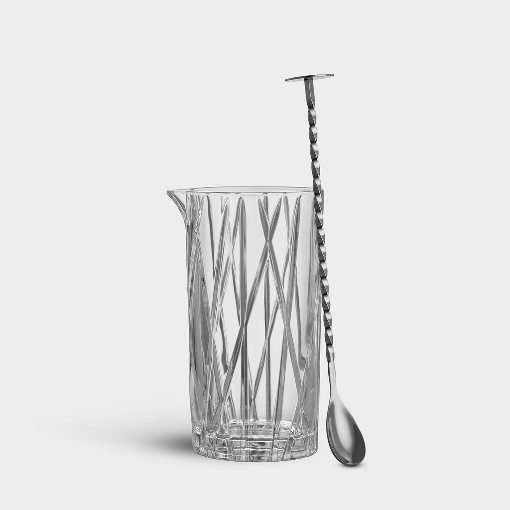 Clear glass pitcher with decorative pattern and a silver spoon on a light gray background