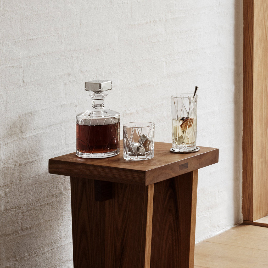 Wooden table with a decanter and glasses against a light brick wall.