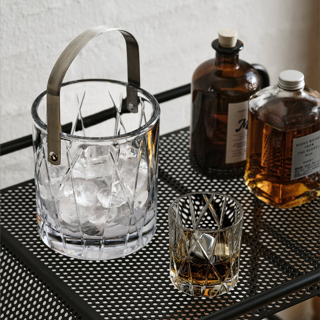 Glass ice bucket with ice cubes, a glass of whiskey, and bottles on a metal tray.