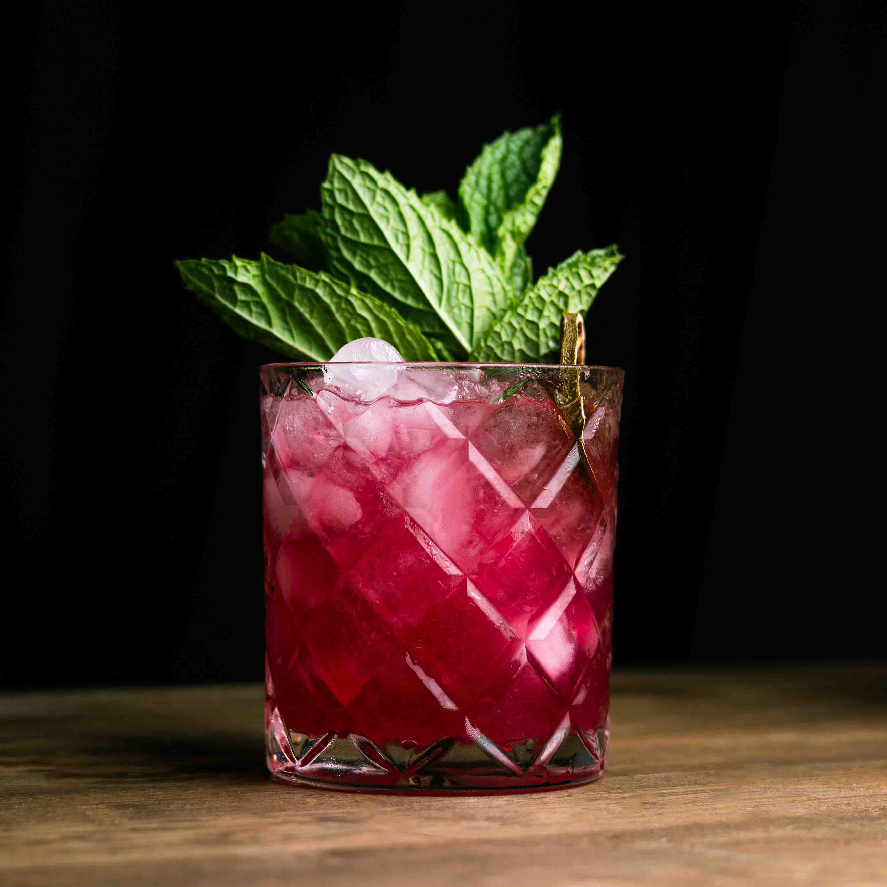 Pink cocktail with ice and mint leaves in a glass on a wooden surface.